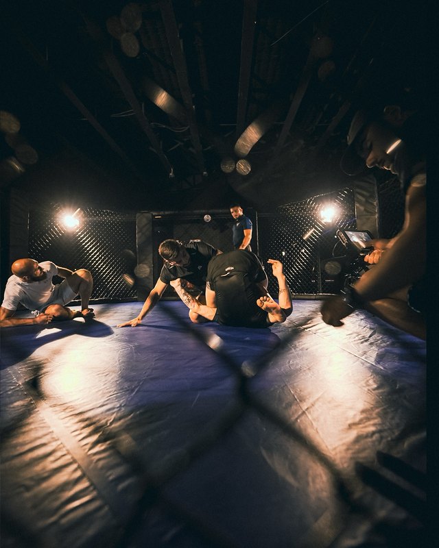 The fight camp seen through the cage: sparring on the blue mat under spotlights.